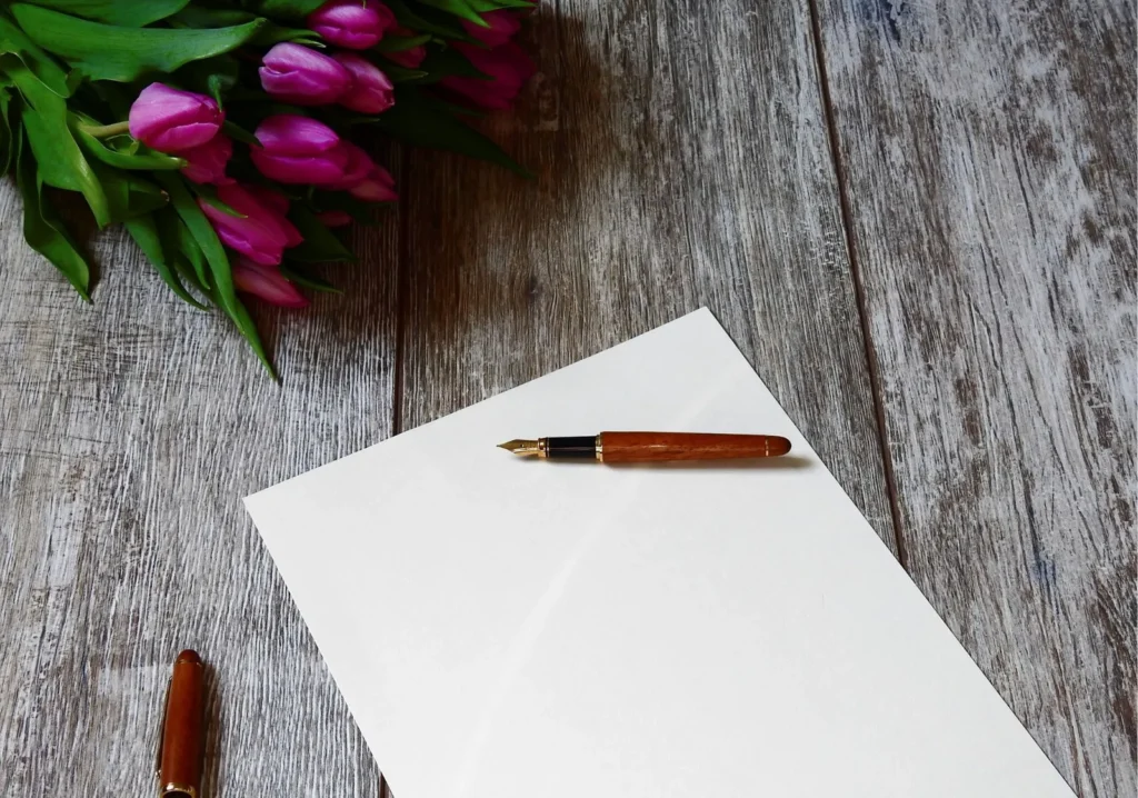 Blank white sheet of paper with pen on it on a gray wooden background with bouquet of purple tulips