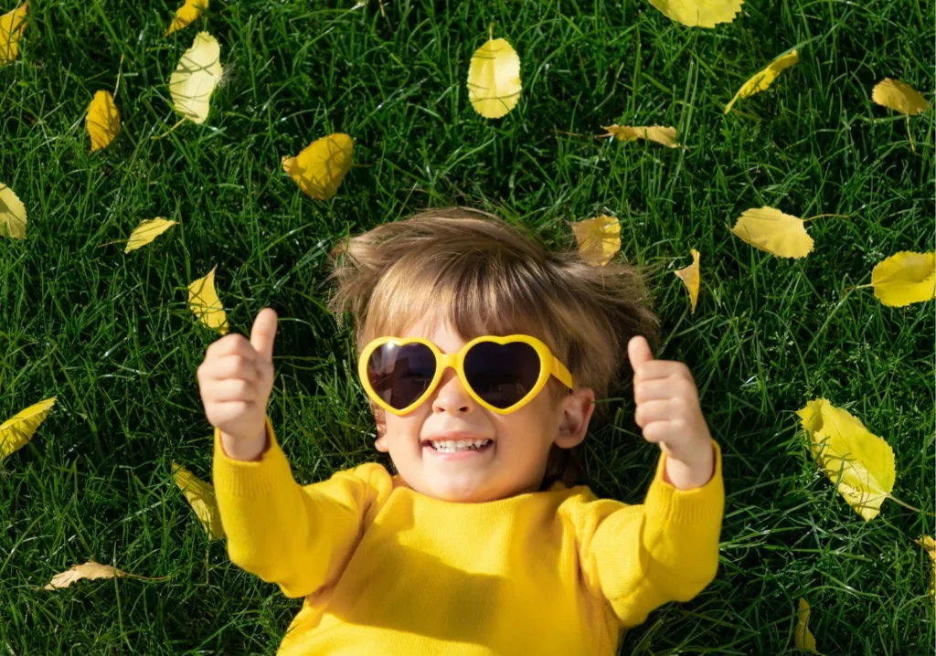 Male toddler laying on grass with yellow heart-shaped sunglasses with 2 thumbs up
