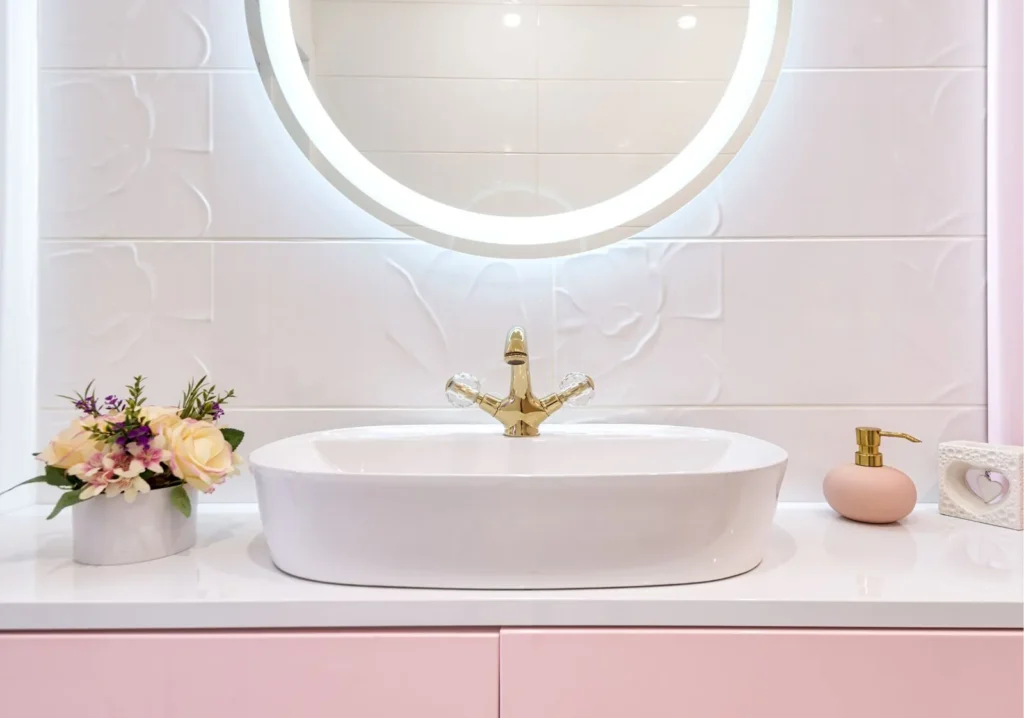 Round vanity mirror with built-in white light over white bathroom sink with pink cabinet