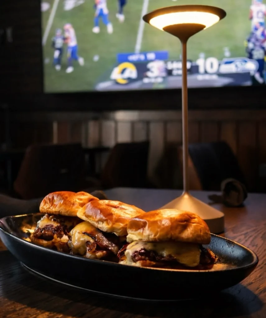 Sliders served under a table lamp at The Tipsy Corner with a game on TV