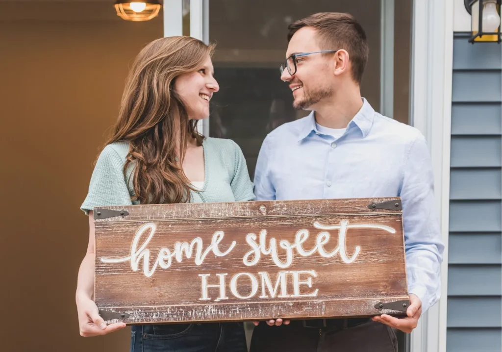 Young couple holding a Home Sweet Home sign on their home's front steps